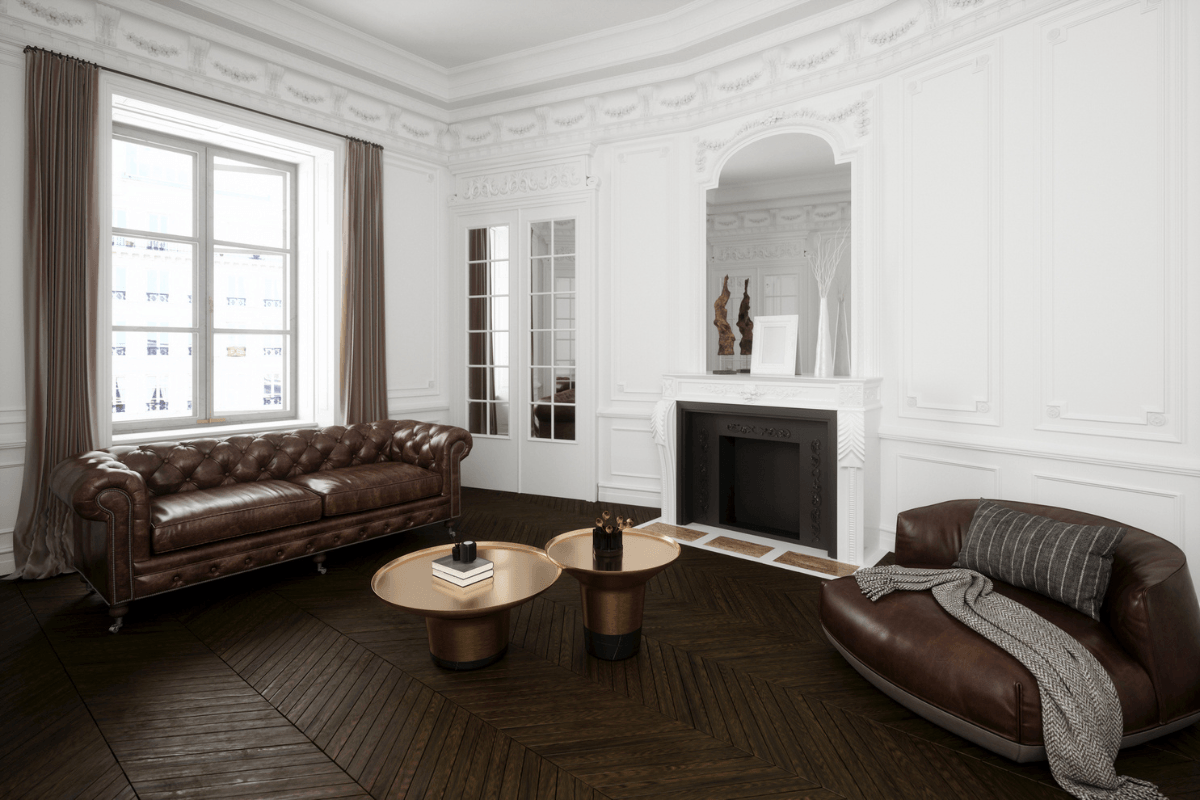 High-ceiling salon exuding Parisian elegance, characterized by white ornamental wall moldings and dark parquet flooring. Prominent features include a vintage-style brown leather Chesterfield sofa, a comfortable leather armchair, and a pair of chic, metallic-based coffee tables.