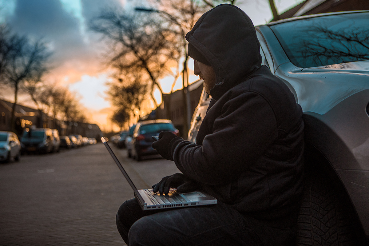 A man in a hoodie sits on the curb beside a parked car, using a laptop, with a sunset in the background