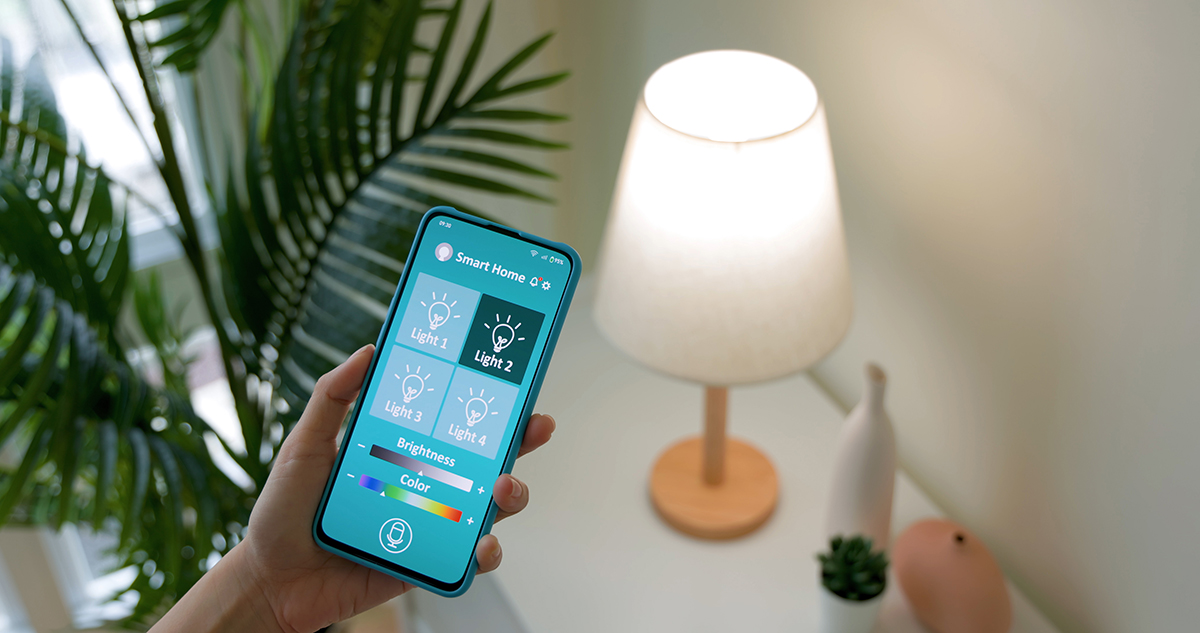 A person holds a mobile phone displaying the smart home app in front of a softly lit table lamp on a white surface.