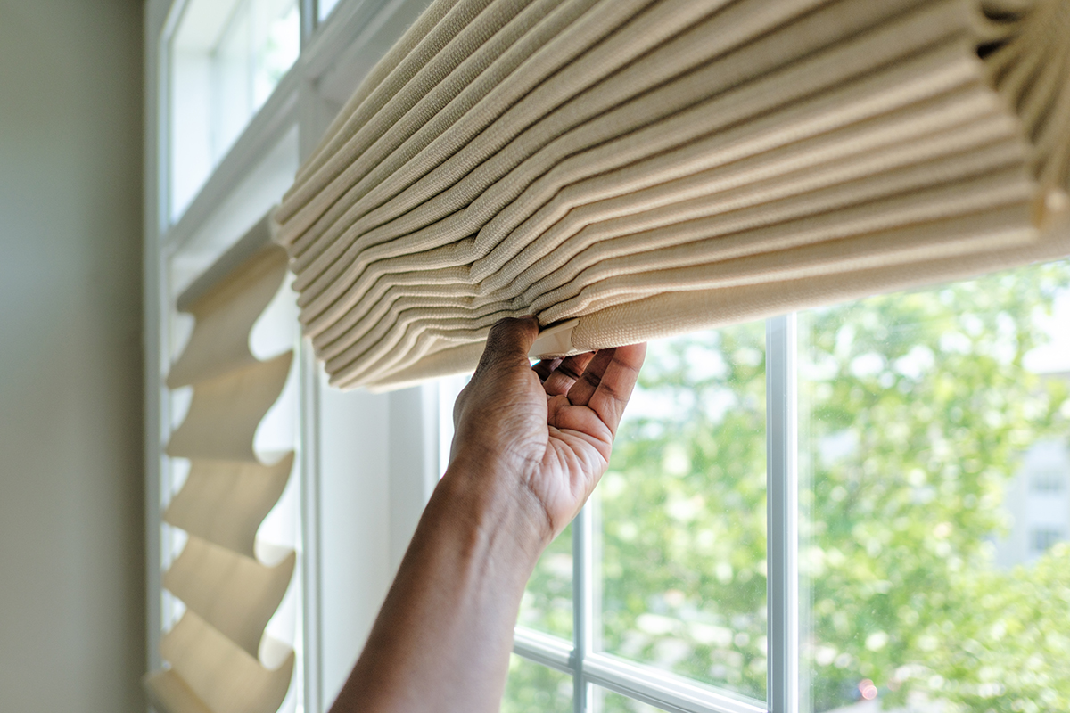 A person pulls down beige pleated blinds over a window, with a view of green plants outside.