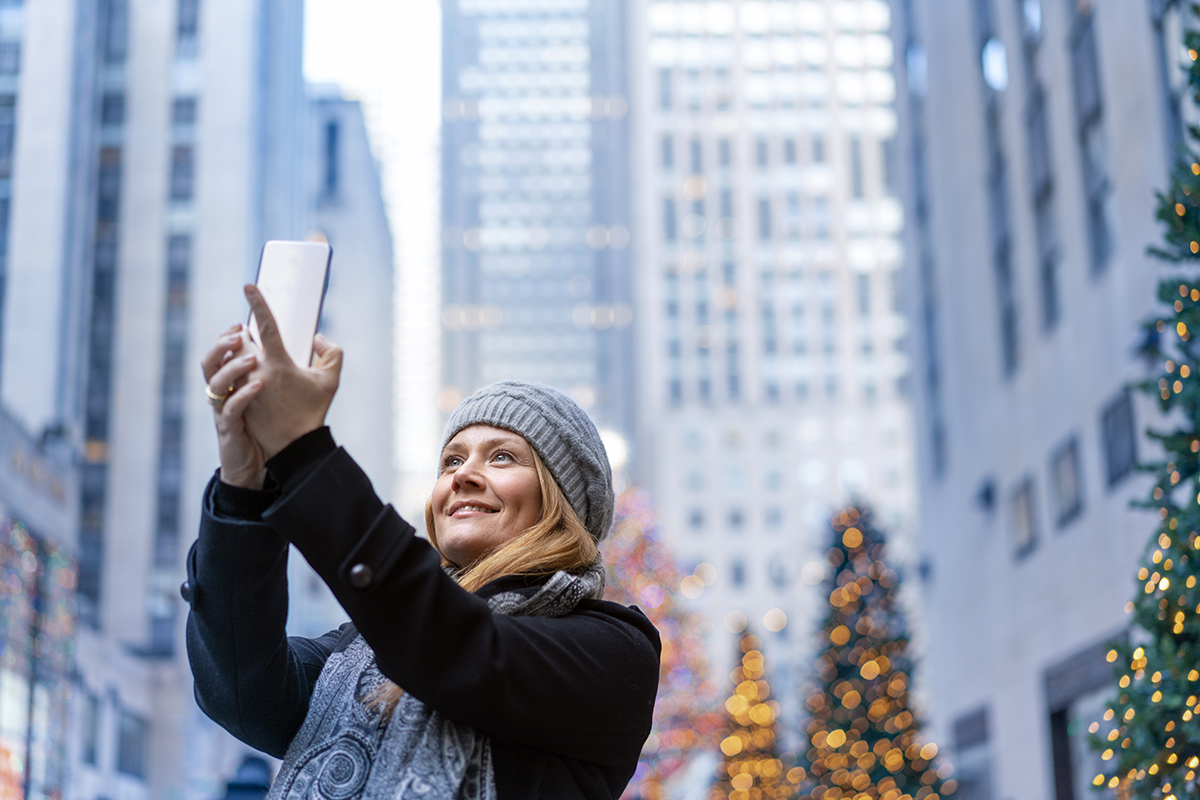 A woman wearing a gray beanie and scarf smiles as she takes a selfie with her smartphone, surrounded by festive Christmas trees and tall city buildings.