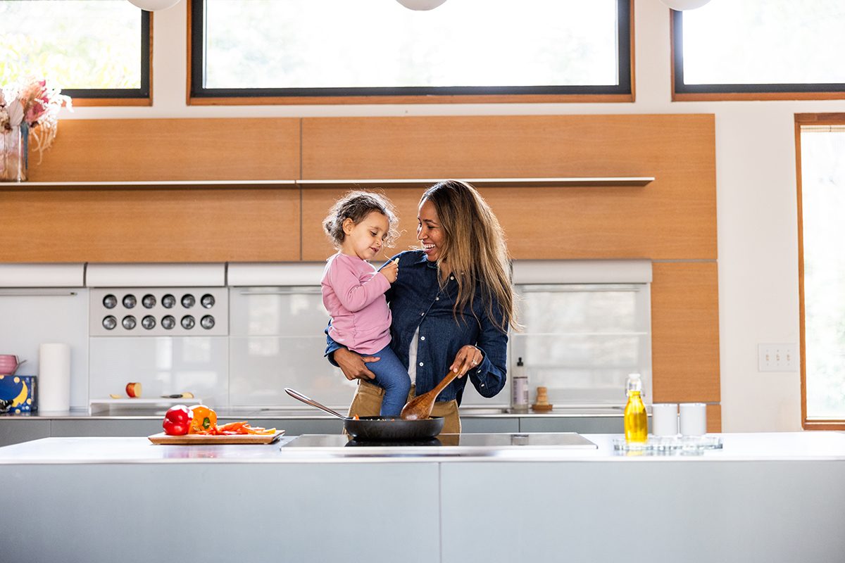 A smiling woman is holding her daughter and cooking in a modern kitchen